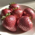 Five fresh strawberries dusted with shimmering rose gold metallic powder, sitting in a white bowl.