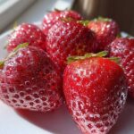 Close-up of vibrant red Rose Gold Metallic Dusted Strawberries glistening on a white plate in bright light.