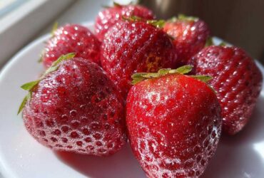 Close-up of vibrant red Rose Gold Metallic Dusted Strawberries glistening on a white plate in bright light.
