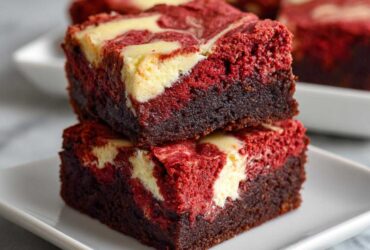 Two stacked, rich Salted Caramel Red Velvet Brownies featuring a dark chocolate base and a vibrant red top layer swirled with cream cheese.