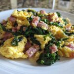 Fluffy scrambled eggs with dark green kale and pink minced ham served on a white plate.