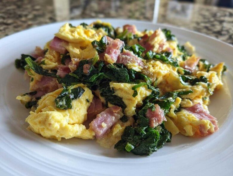 Fluffy scrambled eggs with dark green kale and pink minced ham served on a white plate.