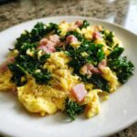 Fluffy scrambled eggs with dark green kale and pink minced ham cubes, seasoned with black pepper.