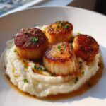Four perfectly seared scallops resting on a bed of creamy cauliflower puree, drizzled with sauce and garnished with chives.