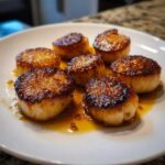 Six perfectly seared scallops glistening in grapefruit brown butter sauce on a white plate.