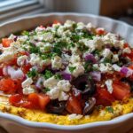 A vibrant close-up of the top layer of the Seven-Layer Mediterranean Dip, featuring feta, tomatoes, olives, and herbs.