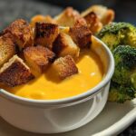 A white bowl filled with bright orange Sharp Cheddar and Apple Cider Fondue, topped with toasted croutons and served with broccoli florets.