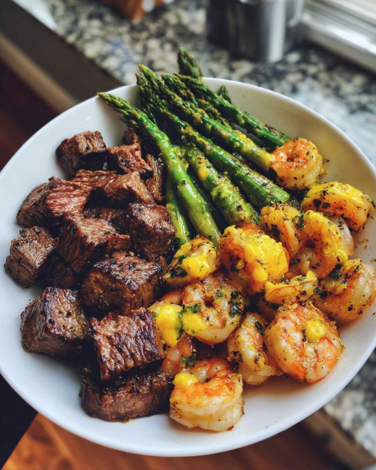 A white bowl filled with chunks of steak, garlic butter shrimp, and roasted asparagus from Sheet Pan Steak and Garlic Butter Shrimp.