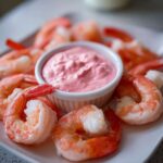 Close-up of cooked shrimp arranged around a small white bowl of bright pink remoulade sauce for Shrimp Cocktail.