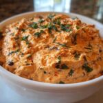 A close-up of creamy Smoked Paprika Neiman Marcus Dip garnished with parsley and paprika in a white bowl.