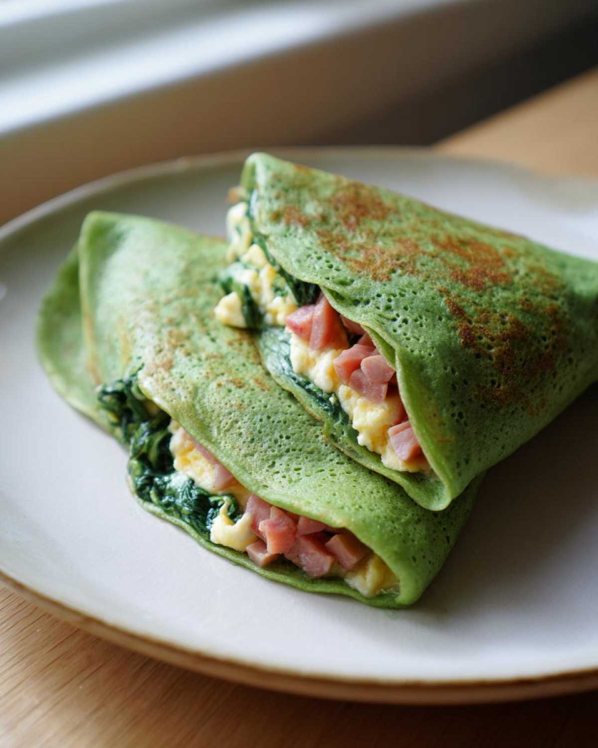 Two folded Spinach Crepes with Ham and Egg Filling stacked on a light plate.