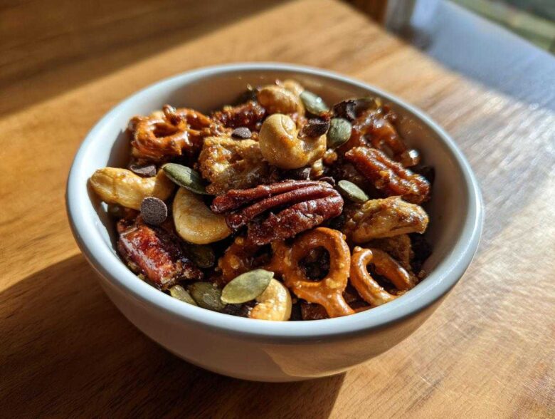 Close-up of a white bowl filled with Sweet and Salty Gameday Trail Mix, featuring pecans, cashews, pretzels, and chocolate chips.