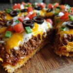 A slice cut from a Taco Pizza with Corn Chip Topping showing seasoned ground beef, melted cheese, tomatoes, olives, and green onions.