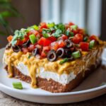 A thick slice of Vegan 7-Layer Dip featuring cashew queso dripping over layers of cream and bean dip, topped with tomatoes and olives.