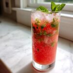 A tall glass filled with a bright red Watermelon Mint Refresher, ice, muddled mint, and garnished with fresh mint sprigs.