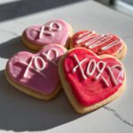 Four heart-shaped XOXO Frosted Sugar Cookies with pink and red icing sitting on a light countertop.