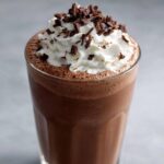 Close-up of a tall glass filled with rich, blended frozen hot chocolate topped with whipped cream and chocolate shavings.