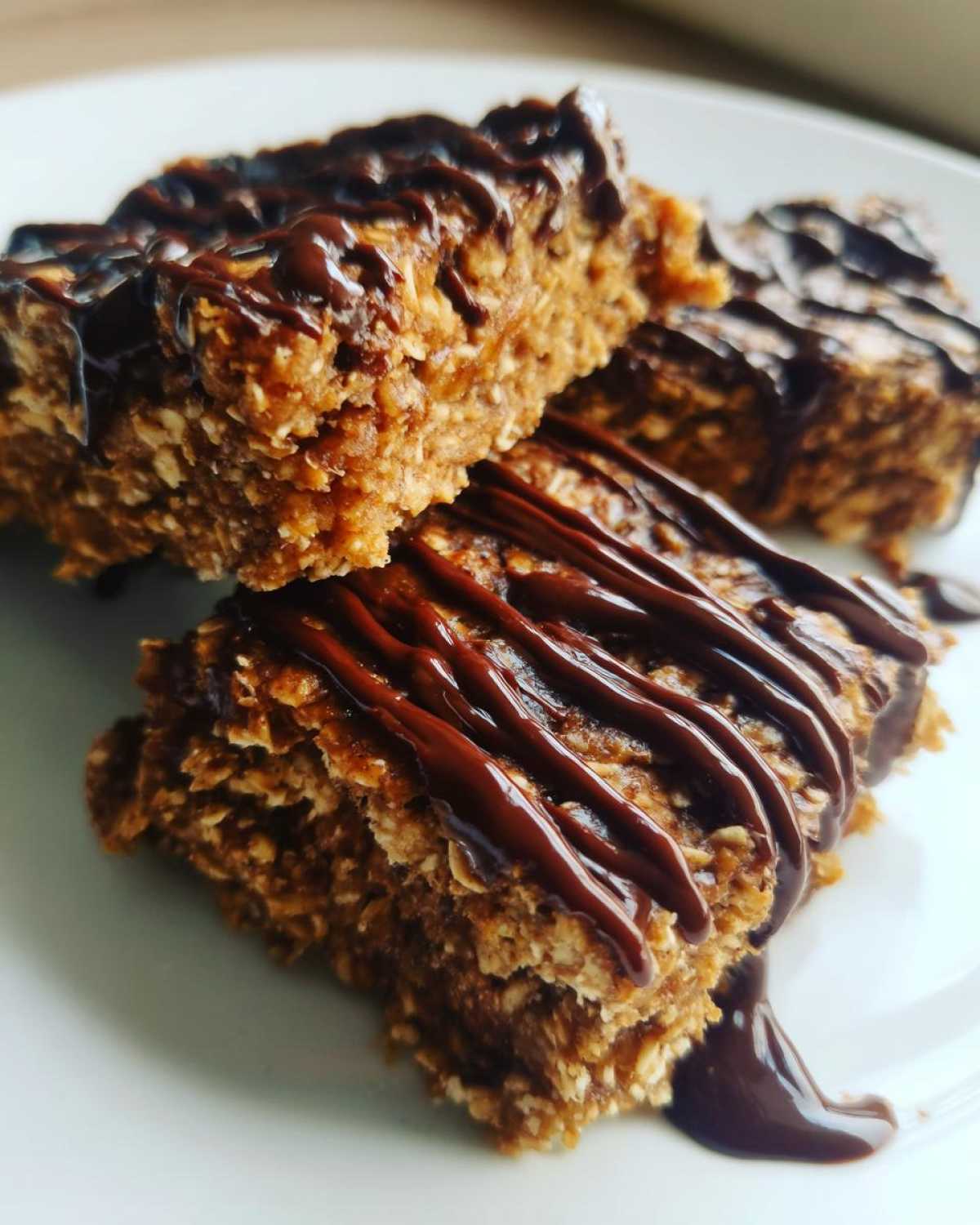 Close-up of three homemade protein bars stacked on a white plate, drizzled generously with melted dark chocolate.