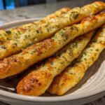 Close-up of several golden brown, herb-topped breadsticks stacked in a light-colored bowl.