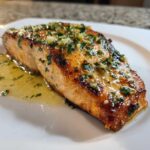 Close-up of a juicy, golden-brown air fryer salmon fillet topped with melted butter, garlic, and fresh parsley.