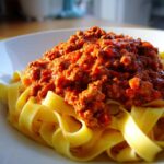 Close-up of rich, meaty bolognese sauce generously spooned over a bed of fresh yellow tagliatelle pasta.