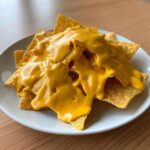 Close-up of tortilla chips completely covered in thick, bright yellow nacho cheese sauce on a white plate.