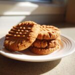 A stack of four golden brown peanut butter cookies with classic fork marks, sitting on a white plate in bright sunlight.