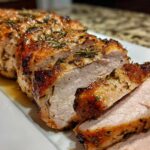 Close-up of a perfectly cooked and sliced pork loin roast seasoned with herbs on a white platter.