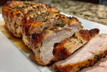 Close-up of a perfectly cooked and sliced pork loin roast seasoned with herbs on a white platter.