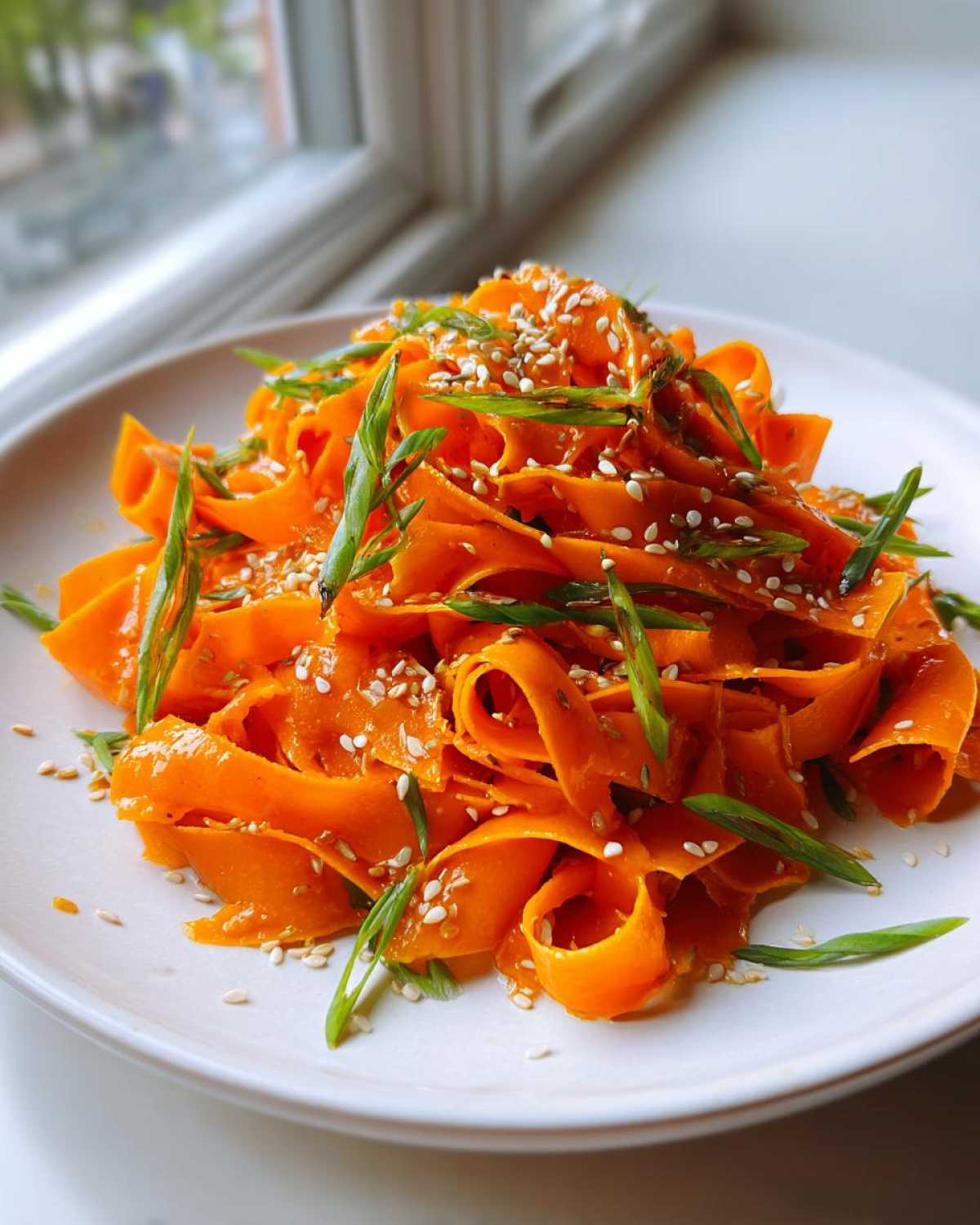 Close-up of vibrant raw carrot salad ribbons tossed in dressing, topped with sesame seeds and sliced green onions.
