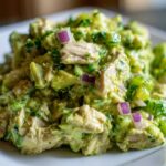 Close-up of creamy avocado chicken salad chunks mixed with red onion and green herbs on a white plate.