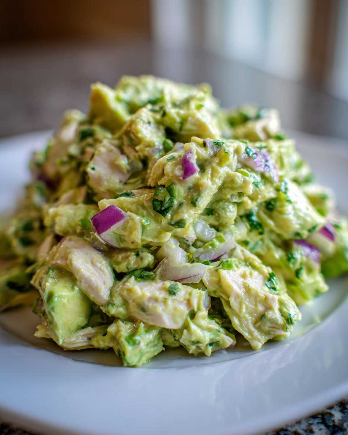 A close-up shot of a generous serving of creamy avocado chicken salad mixed with red onion chunks on a white plate.