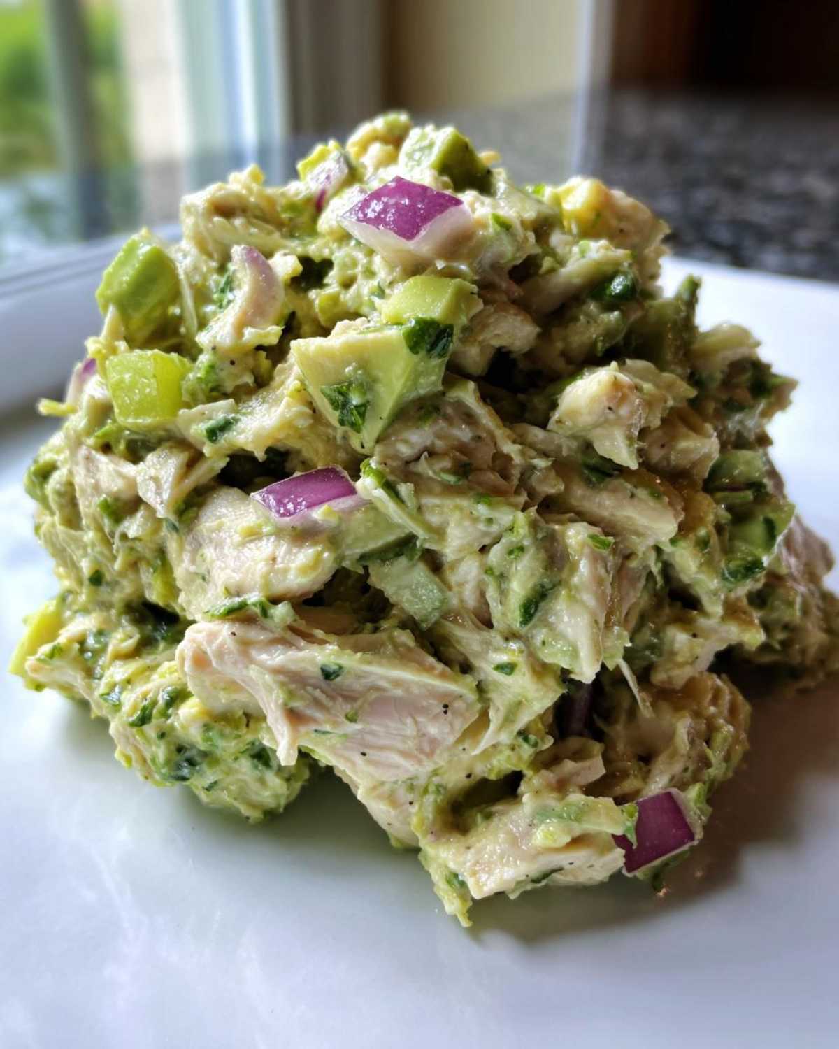 A mound of creamy avocado chicken salad featuring chunks of white meat chicken, diced avocado, and bright purple red onion pieces.