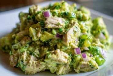 Close-up of creamy avocado chicken salad chunks mixed with red onion and green herbs on a white plate.