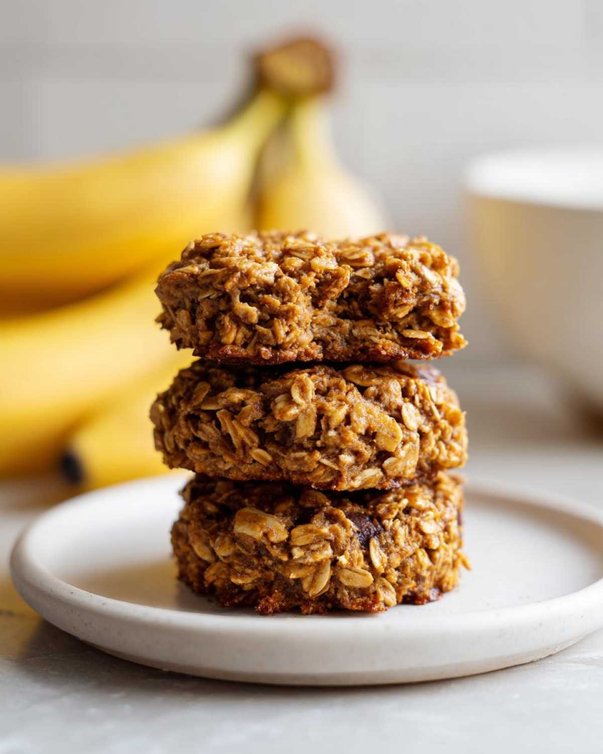 Three stacked banana oatmeal breakfast cookie treats on a small white plate with bananas blurred in the background.