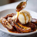 A spoonful of warm caramel sauce is being poured over vanilla ice cream and caramelized bananas in a dish of Bananas Foster.