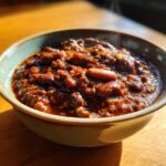Close-up of a steaming bowl filled with rich, dark red chili, featuring beans and ground meat.