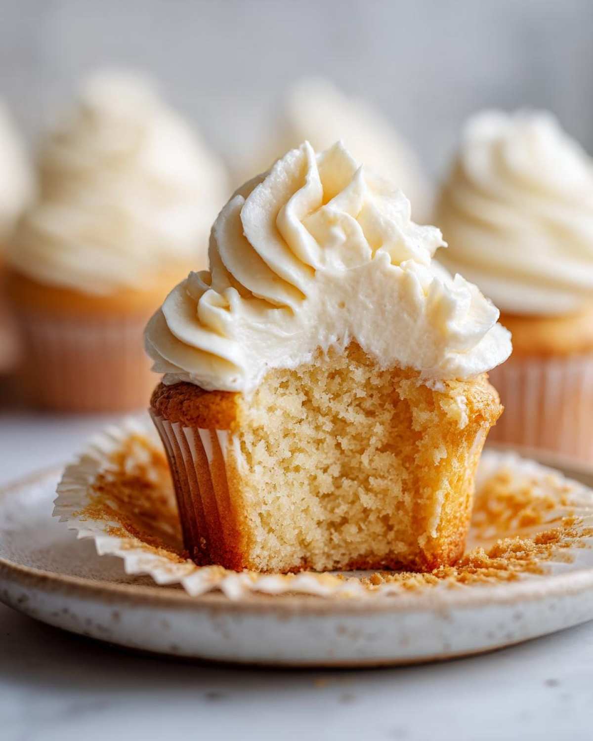 Close-up of a vanilla cupcake with a bite taken out, showing the moist crumb and thick swirl of vanilla frosting.