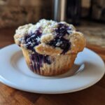A single, freshly baked blueberry protein muffin with a crumbly top sits on a small white plate.