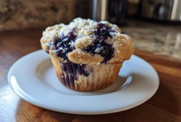 A single, freshly baked blueberry protein muffin with a crumbly top sits on a small white plate.