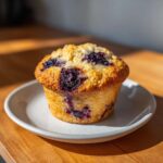 A single, golden-brown blueberry protein muffin with a crumbly top sitting on a small white plate.