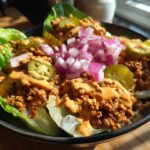 Close-up of a vibrant cheeseburger bowl served in lettuce cups, topped with sauce, pickles, and diced red onion.