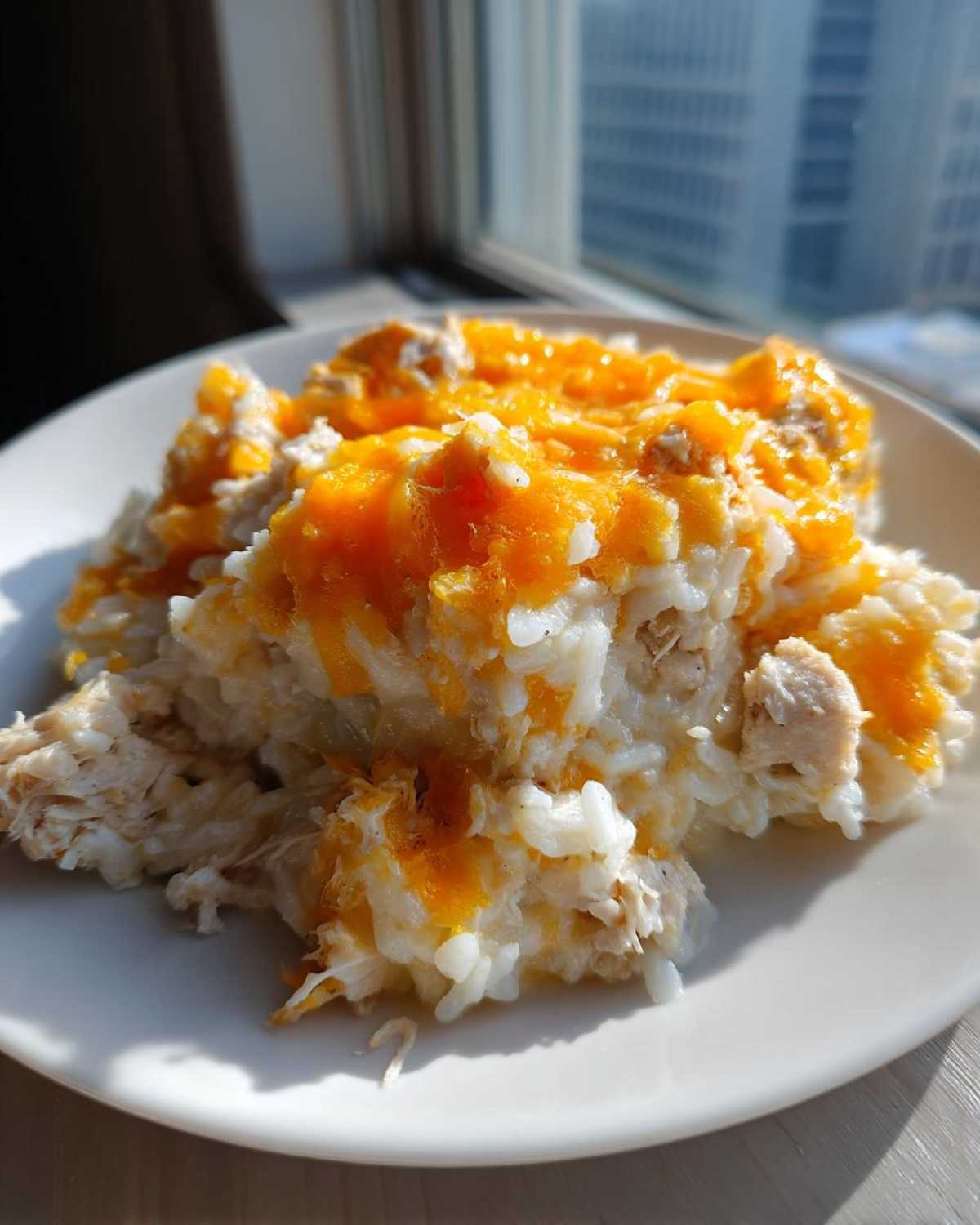 A serving of creamy cheesy chicken and rice topped with melted orange cheddar cheese, photographed near a window.
