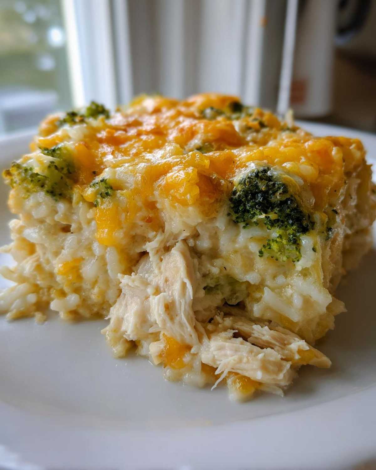 A close-up of a square serving of chicken broccoli rice casserole topped with melted cheddar cheese.