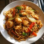 Close-up of a white bowl filled with savory chicken chow mein recipe, featuring noodles, chicken pieces, and colorful vegetables.