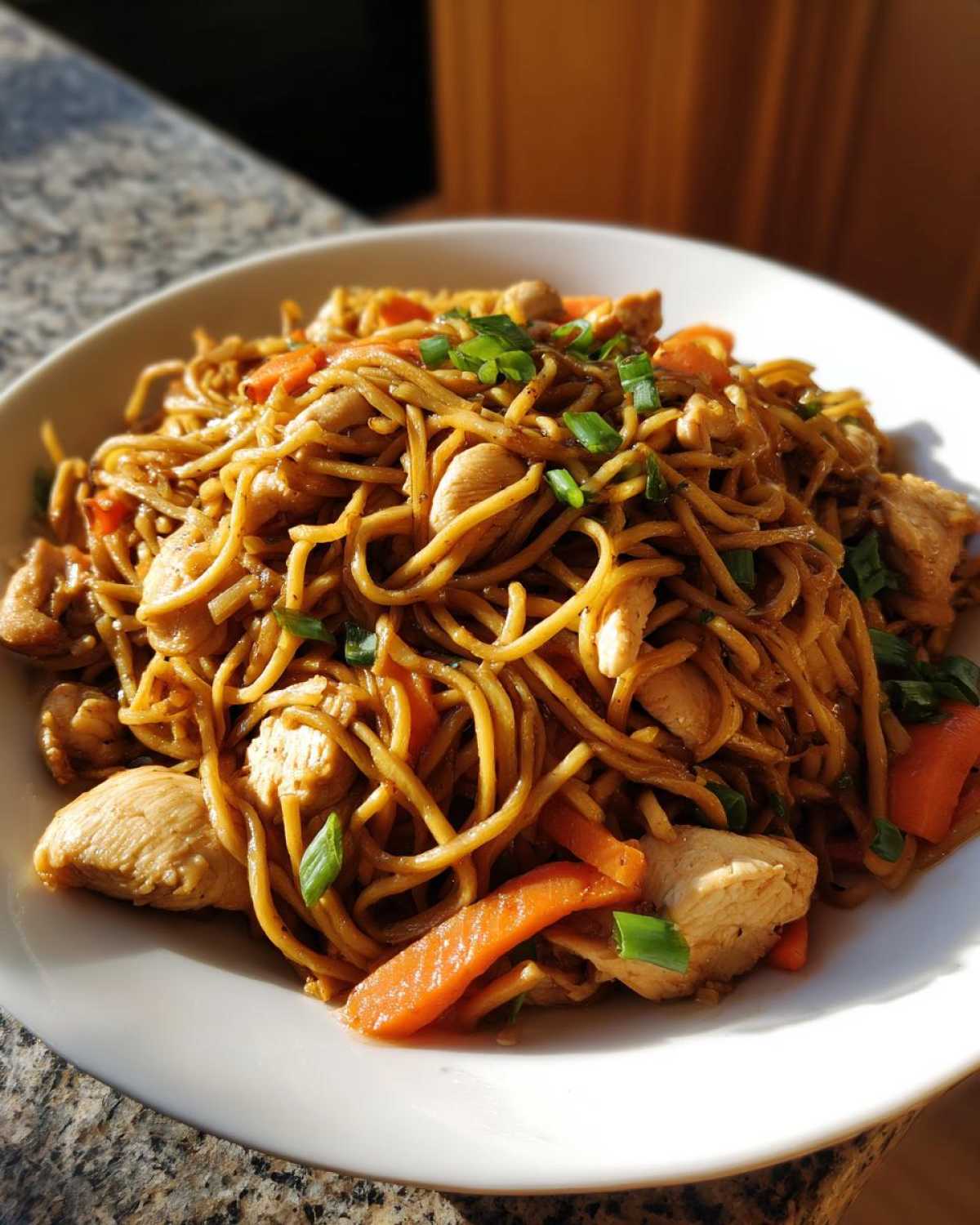 A close-up of a white bowl filled with savory chicken chow mein recipe, featuring noodles, chicken pieces, and carrots, garnished with green onions.