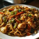 A close-up of a plate piled high with savory chicken chow mein recipe, featuring noodles, chicken chunks, carrots, and green onions.