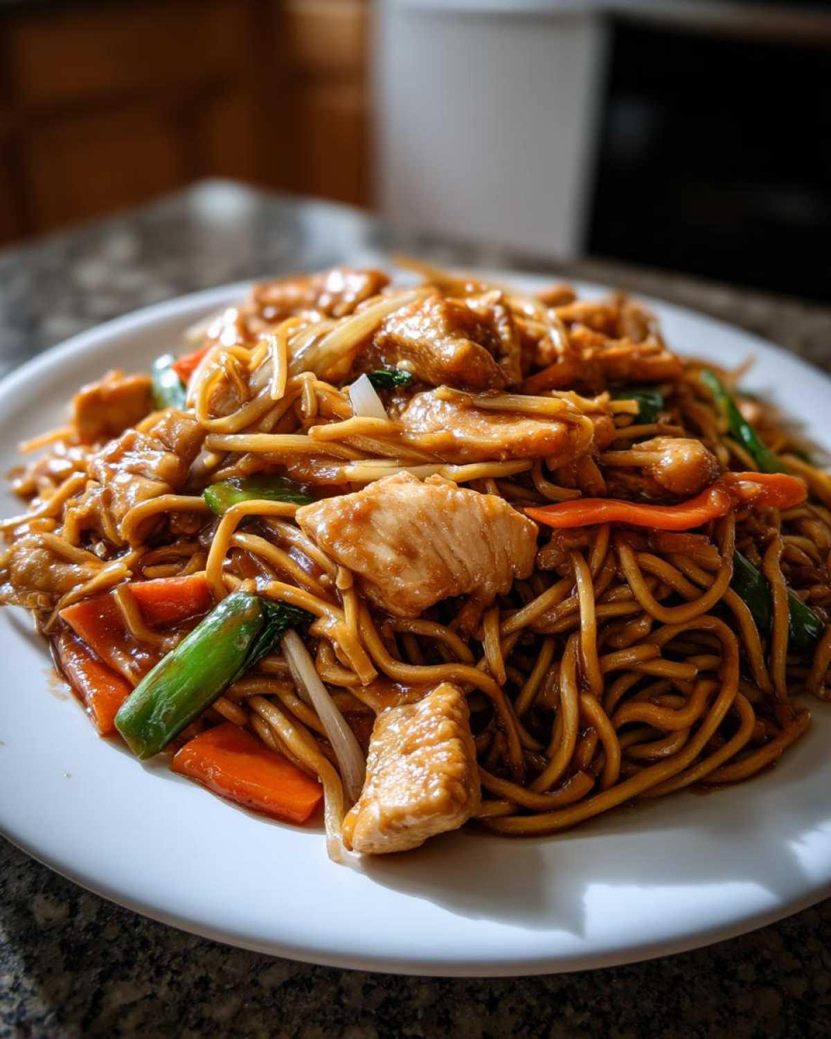 Close-up of a generous serving of chicken chow mein recipe with noodles, chicken pieces, carrots, and green onions on a white plate.