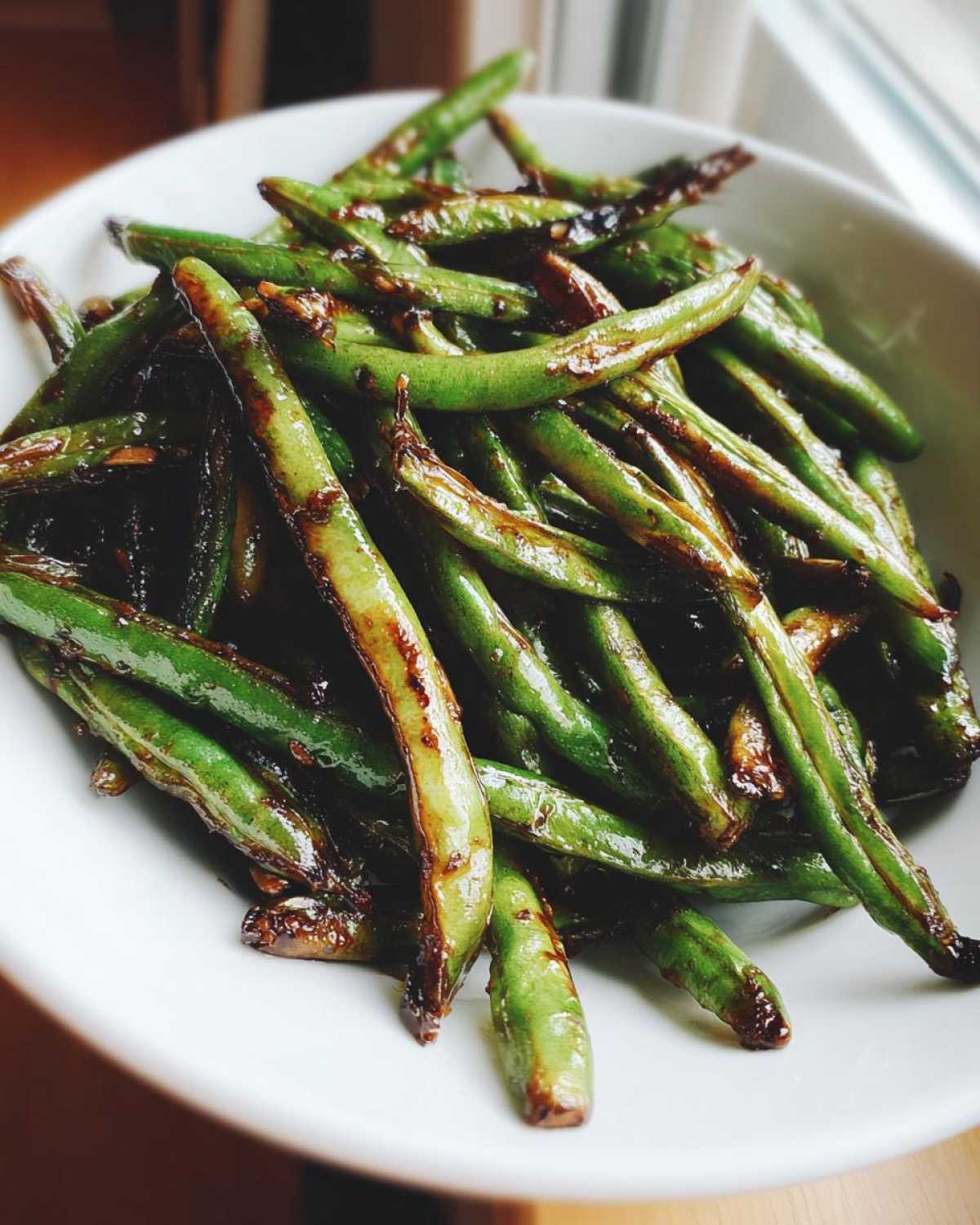 Close-up of vibrant green beans coated in a dark, savory garlic sauce, served in a white bowl.