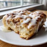 A moist, close-up view of a freshly baked cinnamon roll casserole slice topped with thick white icing.